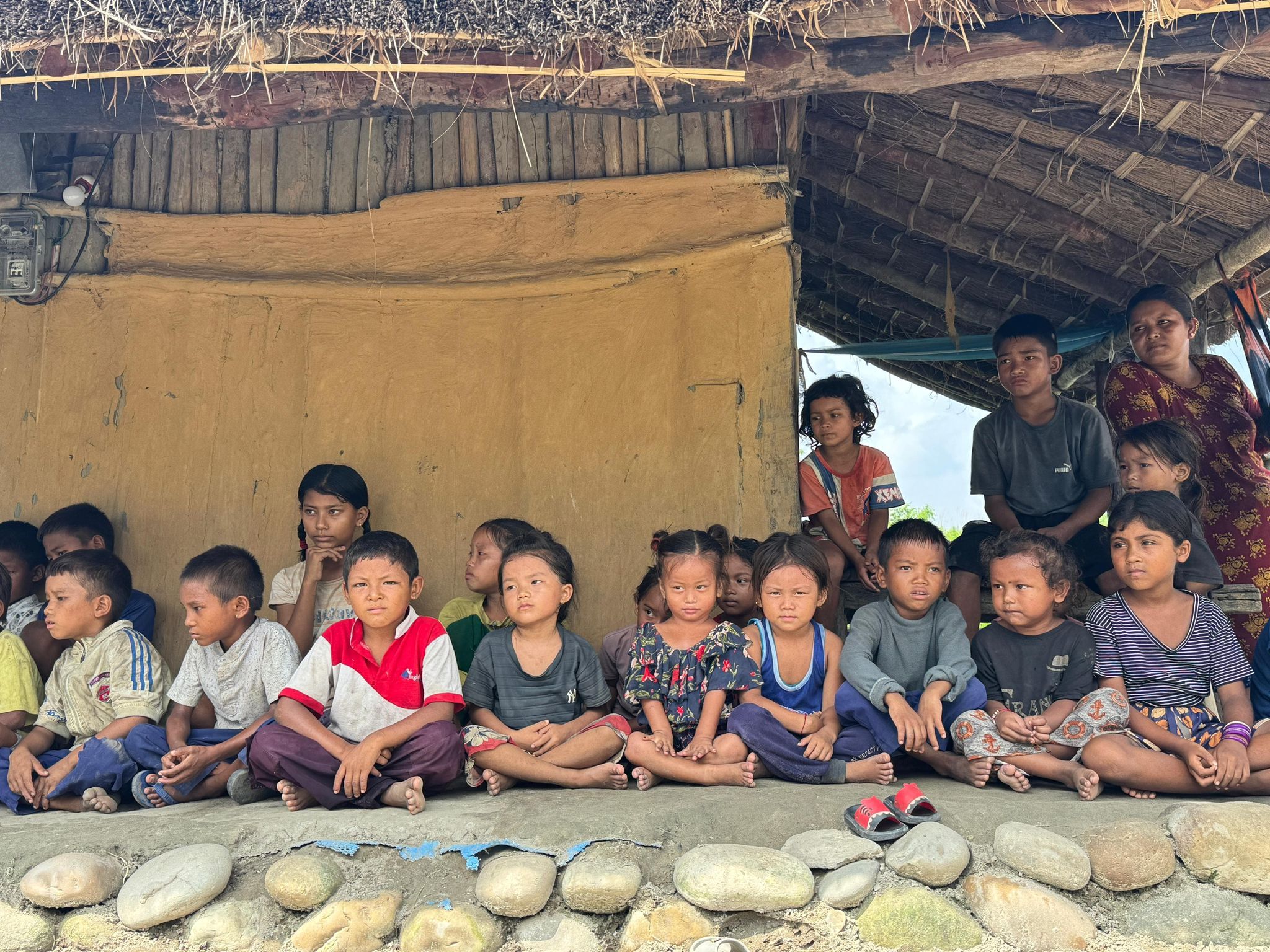 Nepali Children sitting on a stone floor in front of a yellow building with a thatched roof in village in madi valley
